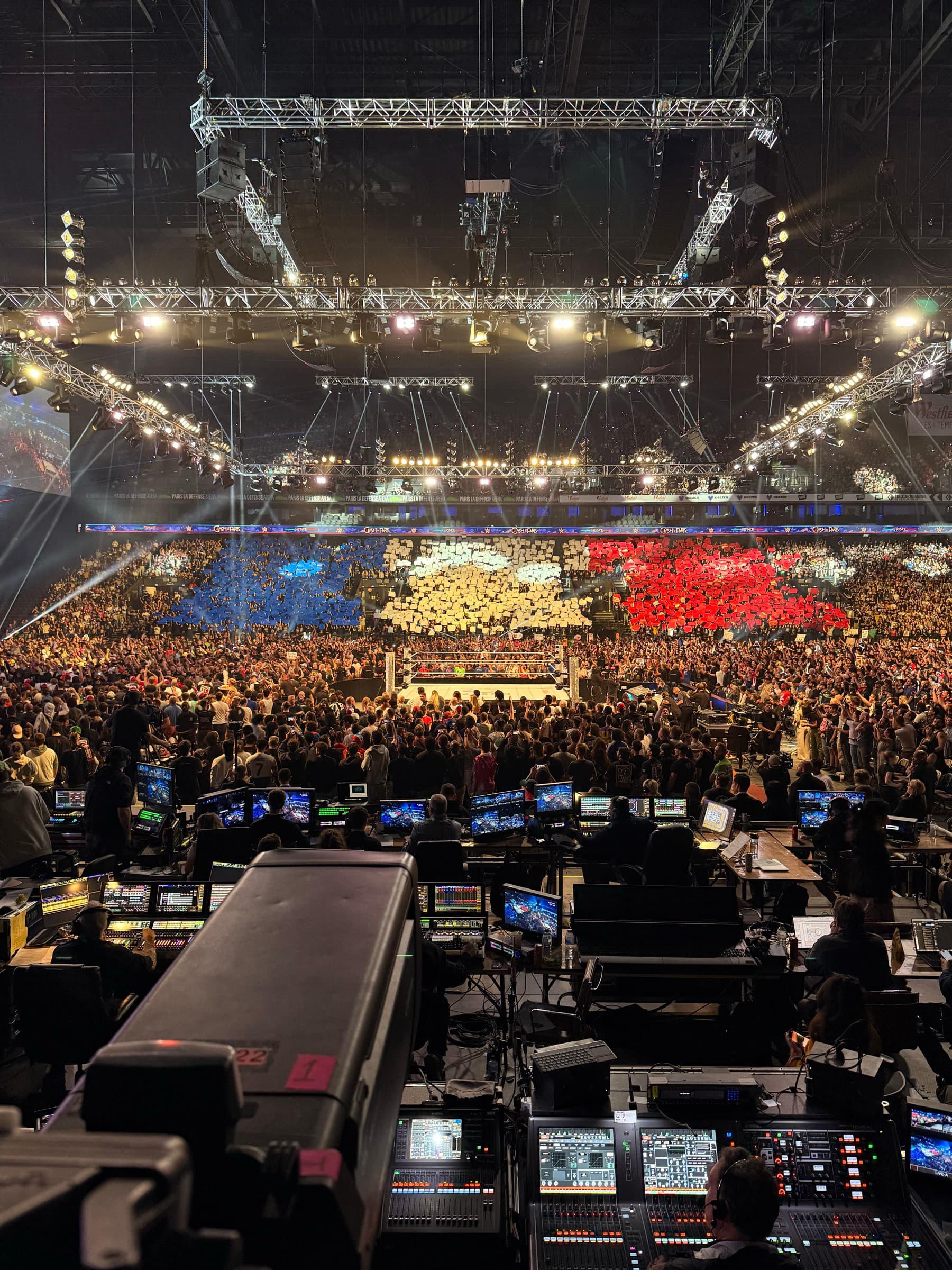 The view from behind the production desk: banks of monitors and mixing gear facing the ring, with the crowd holding up coloured cards